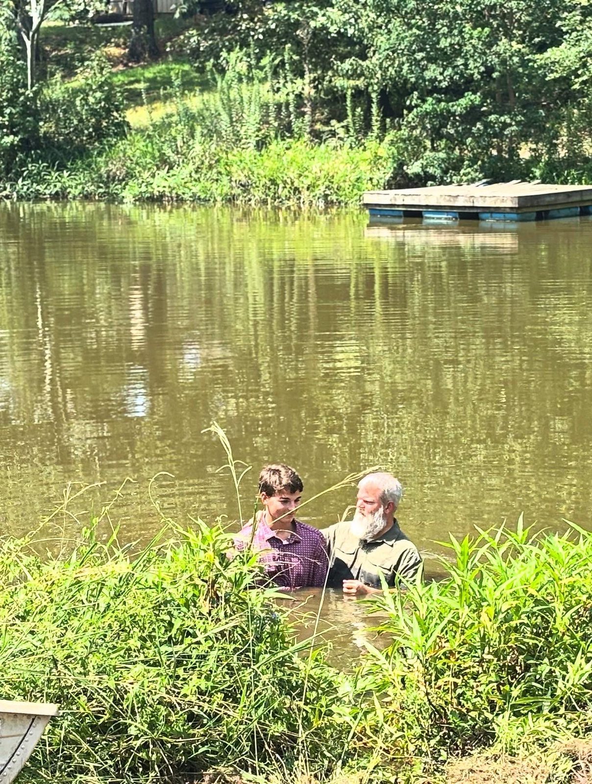 A baptism in the pond, an older man with his arm around a young man, both waist-deep in the water